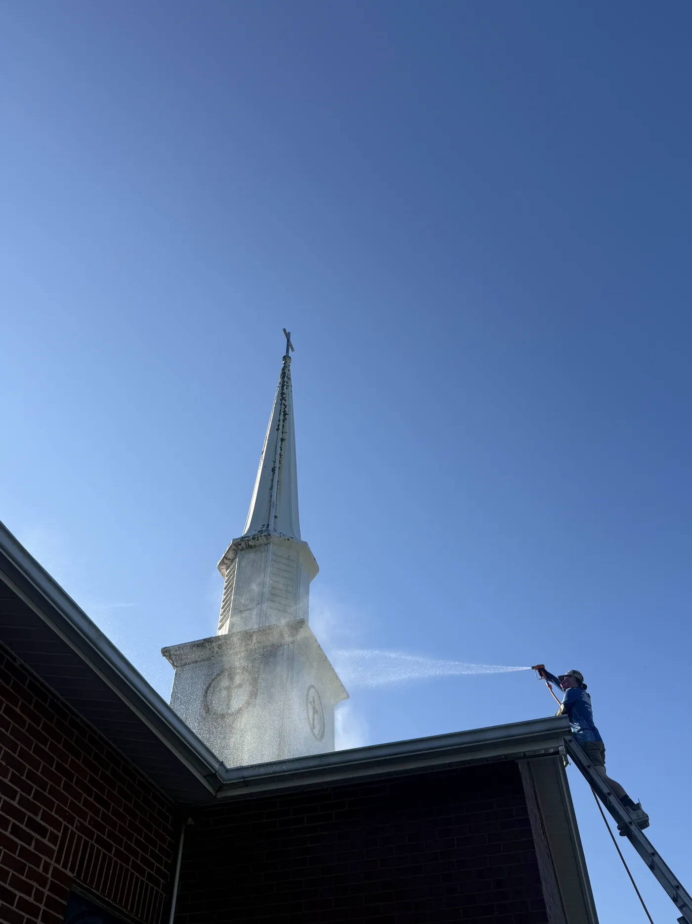 Soft wash spray treating a mold-covered church steeple from an extension ladder