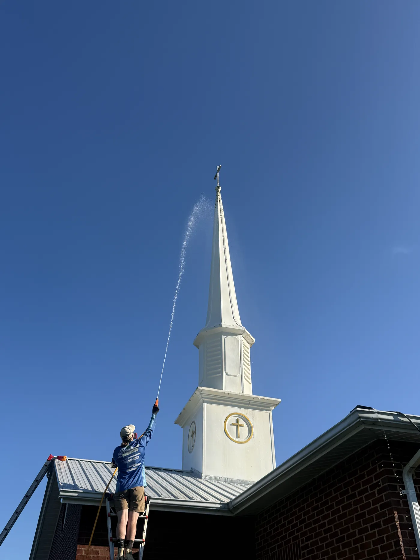 Stand Out technician on a ladder soft washing the upper steeple of a brick church